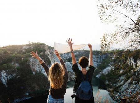 Man Holding White Pennant Beside Girl Raising Two Hands in Front of Lake Surrounded by Mountains