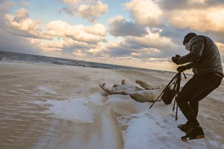 Man in Black Bubble Jacket Walking on Seashore While Holding Black Camera Tripod