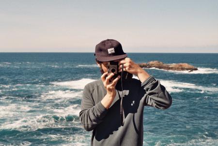 Man in Black Jacket Carrying Point and Shoot Camera in Front of Body of Water during Daytime