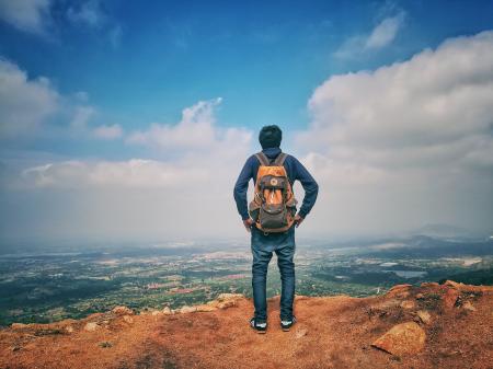 Man in Blue Dress Shirt and Blue Jeans and Orange Backpack Standing on Mountain Cliff Looking at Town Under Blue Sky and White Clouds