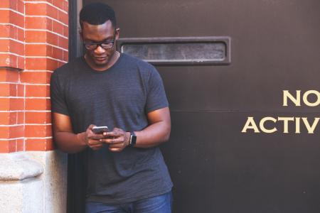 Man in Grey Crew Neck T Shirt Leaning on Brown Brick Wall Holding Smartphone