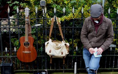 Man Leaning Against Black Steel Fence Beside White and Brown Sling Bag and Brown Acoustic Guitar