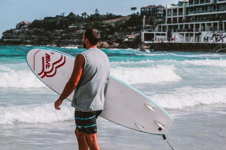 Man Walking Towards Body Of Water Holding A Surfboard