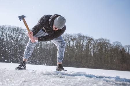 Man Wearing Black Hooded Jacket, Gray Knit Cap, Gray Pants, and Black Shoes Holding Brown Handled Axe While Bending on Snow