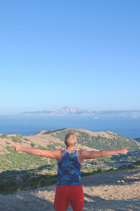 Man Wearing Blue Green and White Tank Top Standing on Brown Mountain Near Blue Body of Water Under Blue Sky during Daytime