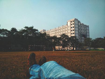 Person in Blue Denim Jean Lying on Brown Grass Field Looking at White Multi-storey Building