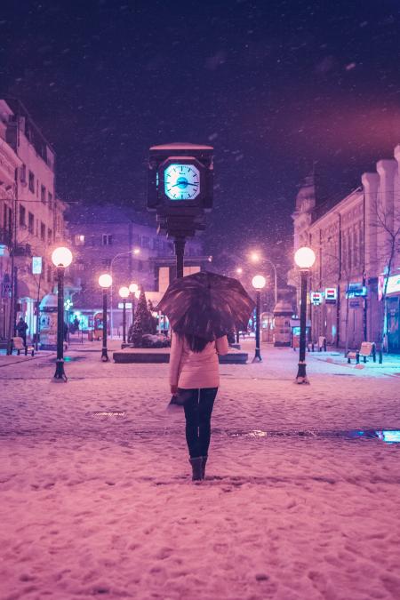 Person in White Long-sleeved Top and Black Pants Under Black Umbrella Standing Near Street Clock