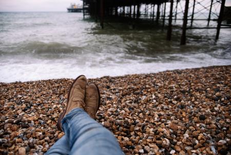 Person Wearing Blue Denim Pants and Brown Boots Sitting on Brown and Black Stone in Front of Body of Water