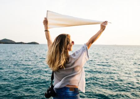 Photo of Woman Wearing White Top, Blue Bottoms and Black Dslr Camera Holding White Textile While Facing the Ocean