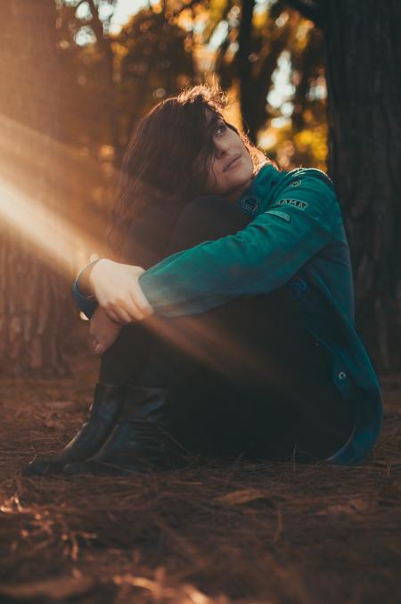 Selective Focus Photography of Woman in Green Jacket and Black Pants Sitting on Field Surrounded With Trees