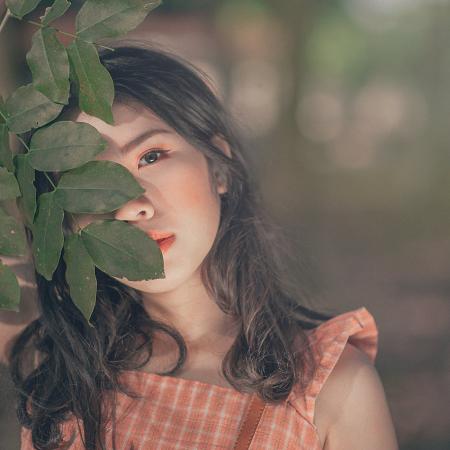 Selective Focus Photography of Woman in Orange Sleeveless Top Hiding Face Behind Tree's Leaf