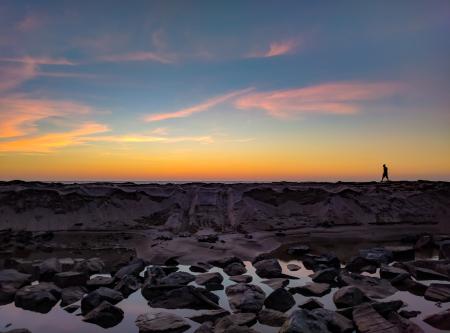 Silhouette Photo of Man Walking on Hill Near Body of Water and Rock Formations