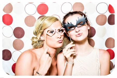 Two Woman Taking Photo in Photobooth Holding Black and Pink Masquerade Mask