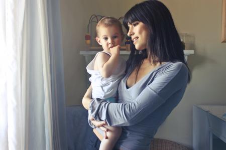 Woman Carrying Baby Boy Wearing White Tank Top Infront of White Curtain Inside the Room