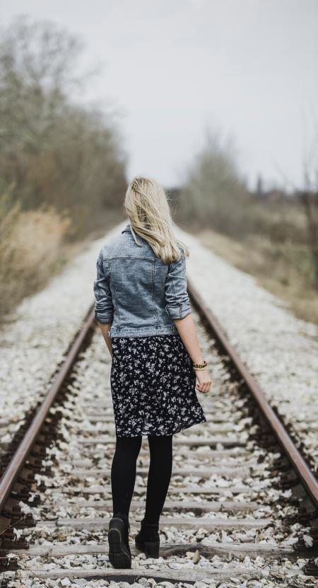 Woman in Blue Denim Jacket, Black Leggings and Black and White Dress Walking on Train Rail