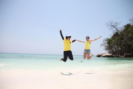 Woman in Blue Denim Mini Short Smiling While Holding Another Person in a Jump Shot Photo at Seashore during Daytime