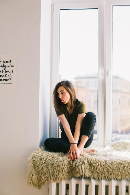 Woman in Green Shirt and Black Denim Jeans Sitting Near Window Inside Room