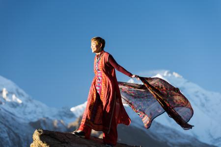 Woman in Orange Dress Standing on Top of Rock Cliff Holding Scarf Near Mountain Covered With Snow
