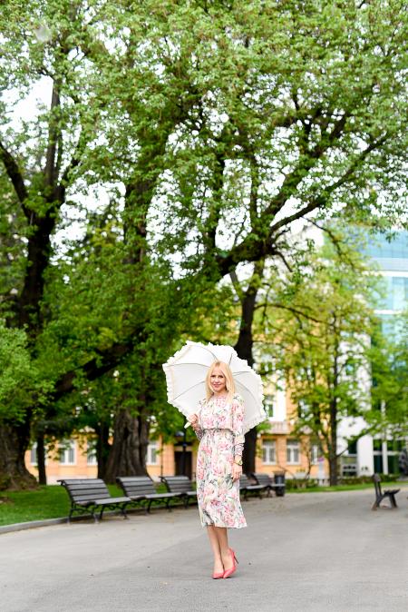 Woman in White Green and Red Floral Holding White Umbrella on Gray Concrete Pathway Near Brown Wooden Bench Near Trees during Daytime