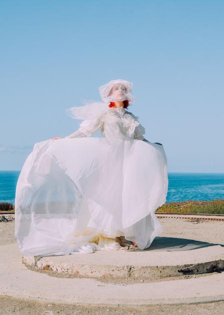 Woman in White Lace Wedding Dress Standing on Gray Pavement Near Body of Water during Daytime