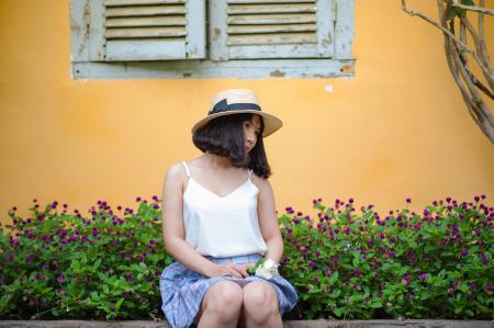 Woman in White Spaghetti Strapped Top With Blue and Purple Plaid Skirt Sitting on Ledge Near Purple Petaled Flower Garden Posing for Photo