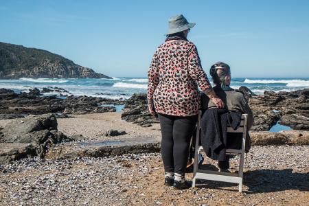 Woman Standing Beside Woman on White Wooden Chair Facing Body of Water