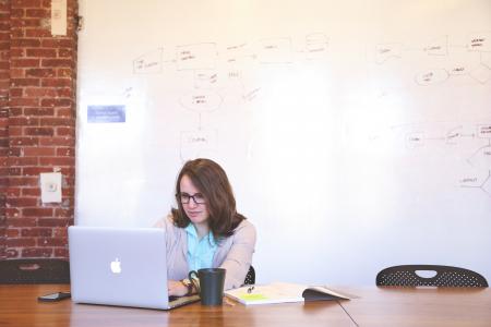 Woman Wearing Black Framed Eyeglasses and Teal Button-up Shirt and Beige Blazer Sitting at Table Near White Macbook