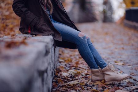 Woman Wearing Black Jacket Blue Distressed Jeans and Brown Boots Sitting on Gray Concrete Barrier