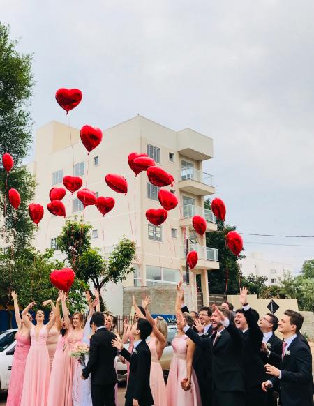 Women Wearing Pink Dresses and Men Wearing Black Suit Jacket and Pants Raising Hands With Red Heart Balloons