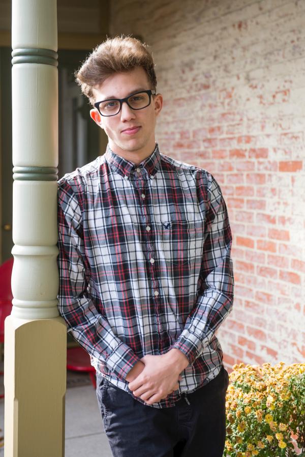 Blonde Man Wearing White, Gray, and Red Plaid Sport Shirt and Black Bottoms Standing While Leaning on Post