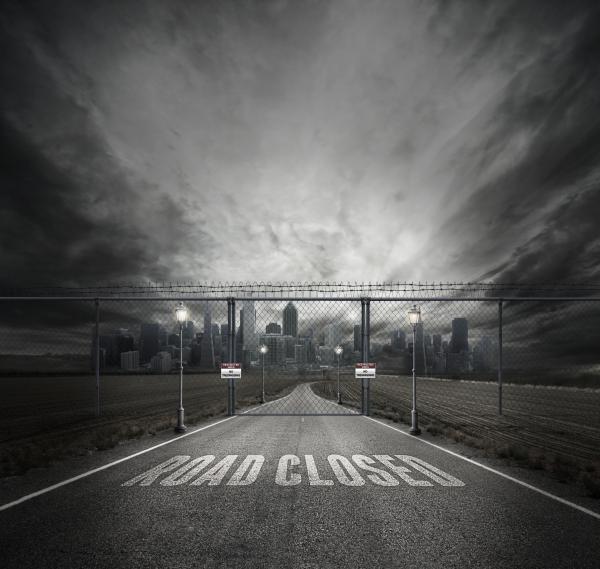 Grayscale Photo of Road Closed on Roadway With Gray Fence Gate in Front of City