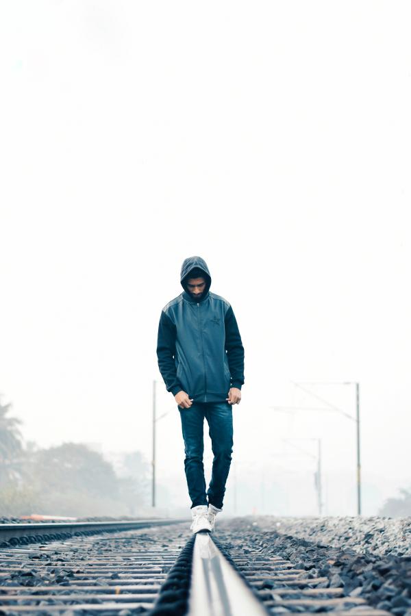 Man Wearing Gray and Black Zip-up Hoodie With Black Denim Jeans and White Shoes Walking on Train Railing Behind White Fog
