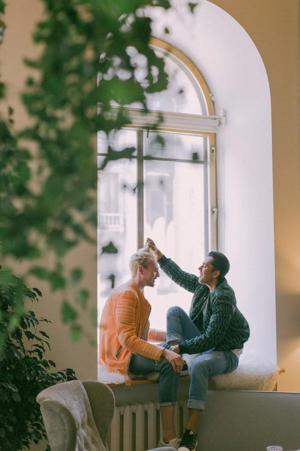 Man Wearing Green Jacket in Front of Man Wearing Orange Zip-up Jacket Sitting on Near the Window Photo