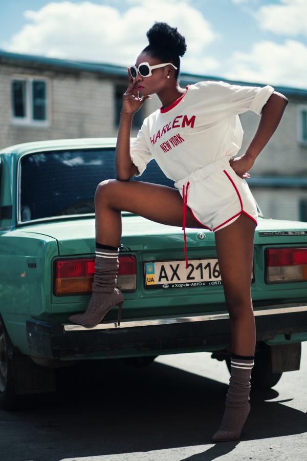 Photo of Woman Wearing White Long-sleeved Shirt and White Dolphin Shorts With Red Trim Standing Near Teal Car