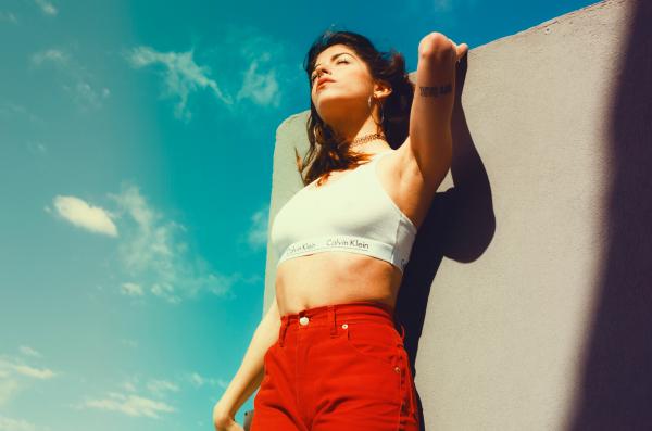 Woman in White Calvin Klein Sports Bra and Red Bottoms Leaning on Gray Wall Under Gray Sky
