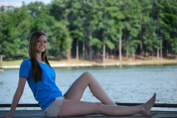 Woman Wearing Blue T Shirt and Beige Short Sitting on Brown Wooden Panel Near Body of Water during Daytime